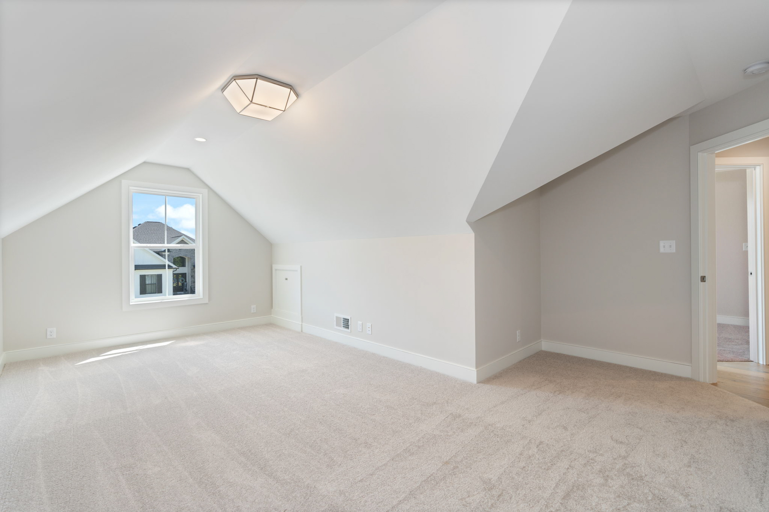 Empty attic bedroom with sloped white ceilings, beige carpet, and a window showing houses outside.