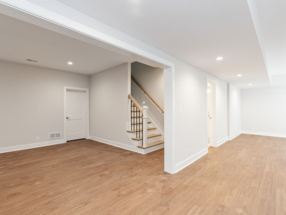 Empty modern basement hallway with stairs on the left, white walls, and recessed ceiling lights.
