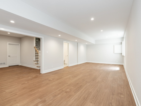 Empty finished basement with light gray walls, warm wooden flooring, and recessed ceiling lights; stairs with a wooden handrail visible on the left side.