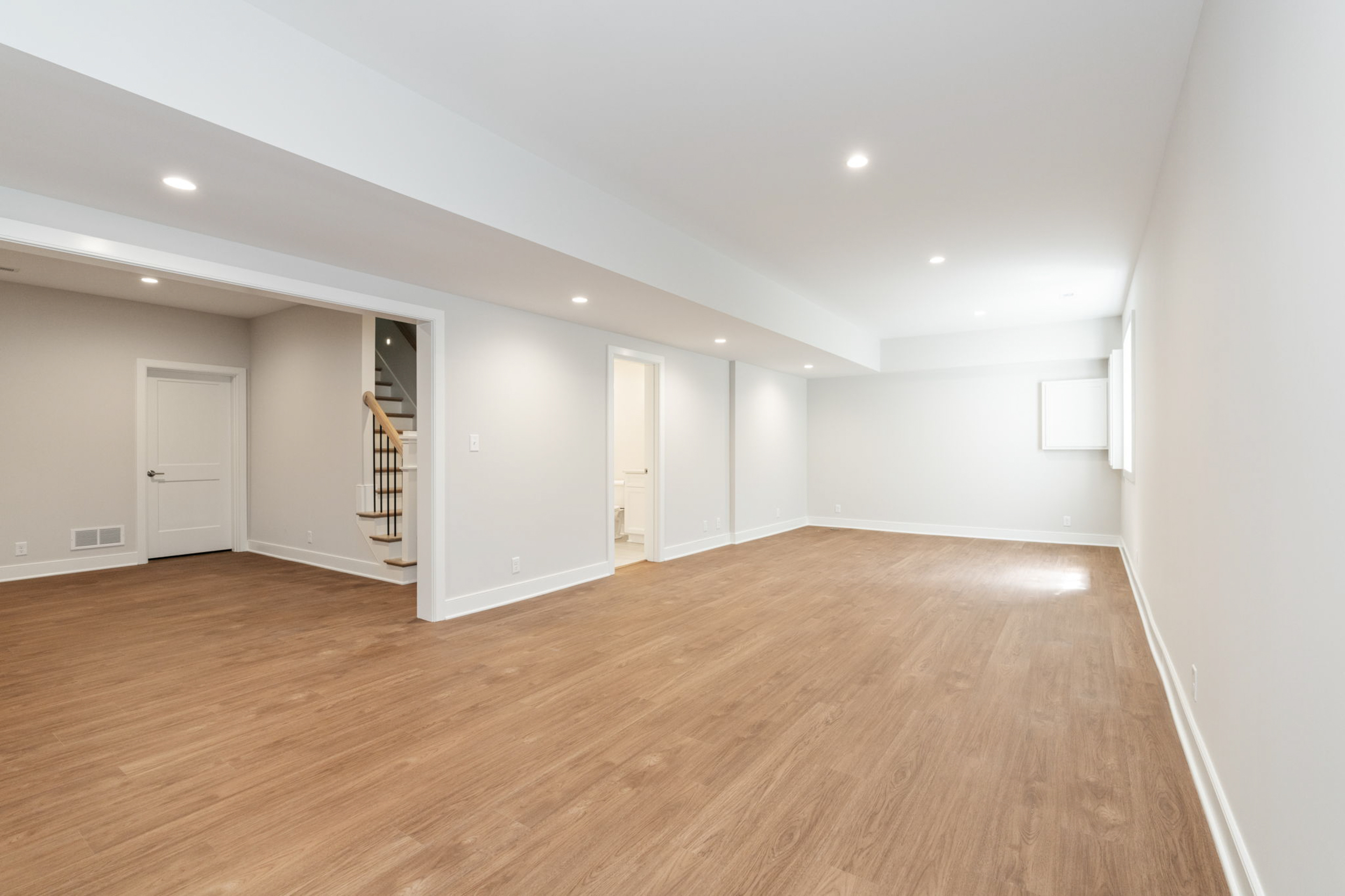 Empty finished basement with light gray walls, warm wooden flooring, and recessed ceiling lights; stairs with a wooden handrail visible on the left side.
