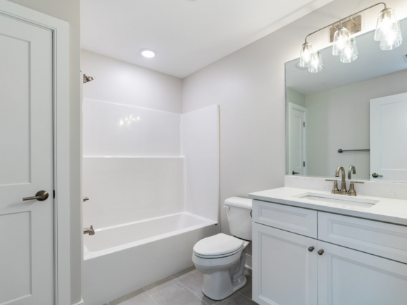 Bright white bathroom with bathtub/shower, toilet, and a white vanity with marble countertop and large mirror in front of sink.