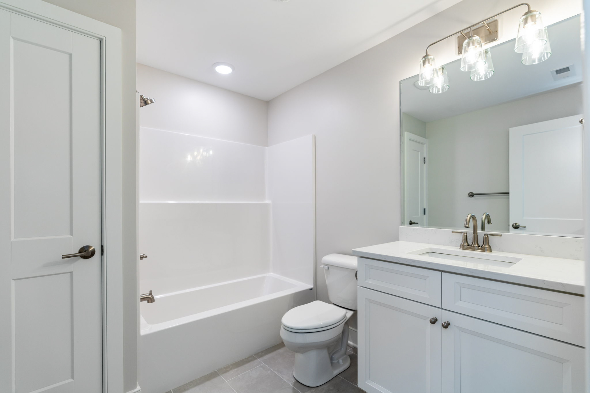 Bright white bathroom with bathtub/shower, toilet, and a white vanity with marble countertop and large mirror in front of sink.