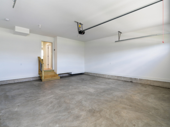 Empty residential garage with concrete floor, white walls, and ceiling-mounted garage door hardware; small wooden stairs lead to an interior door.
