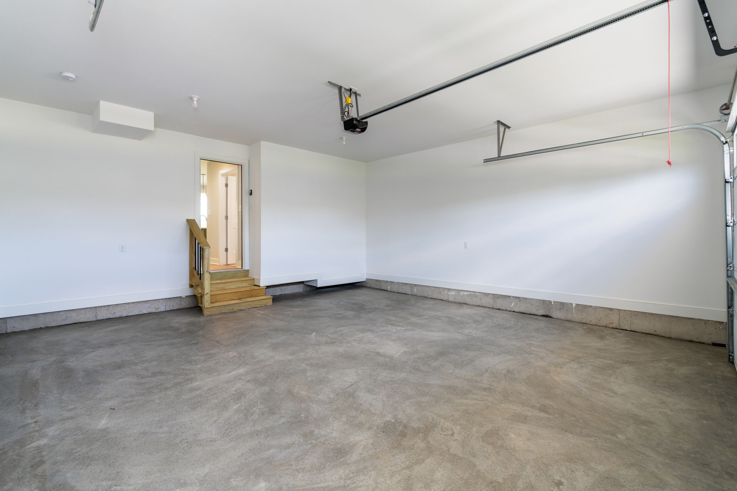 Empty residential garage with concrete floor, white walls, and ceiling-mounted garage door hardware; small wooden stairs lead to an interior door.