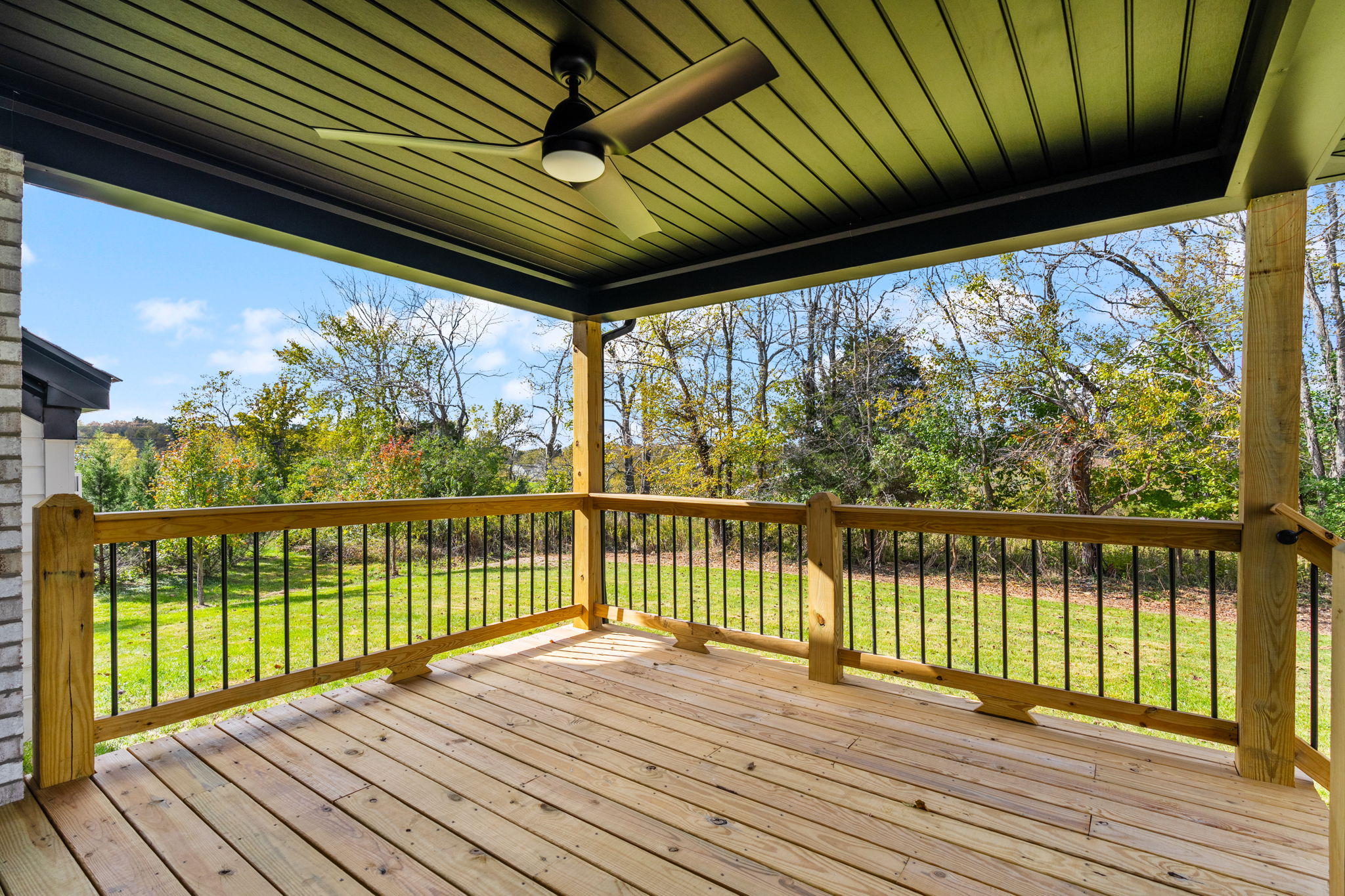 Wood deck under a covered patio with a modern ceiling fan and wood railing, overlooking a green yard and trees.