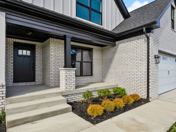 Brick house entry with black front door, brick columns, and concrete steps under an overhang; Modern two-story home with a light brick exterior and garage door nearby.