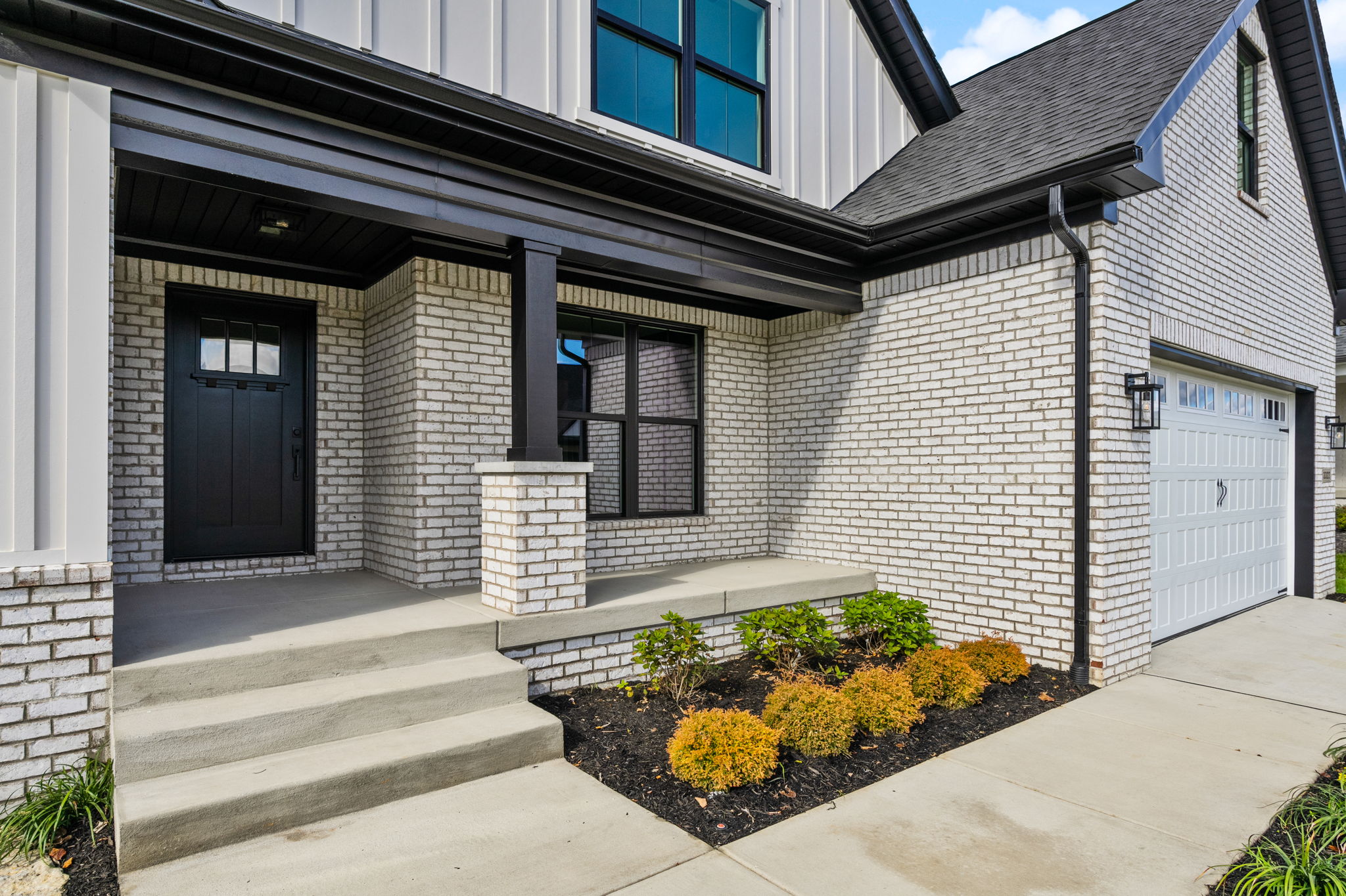 Brick house entry with black front door, brick columns, and concrete steps under an overhang; Modern two-story home with a light brick exterior and garage door nearby.