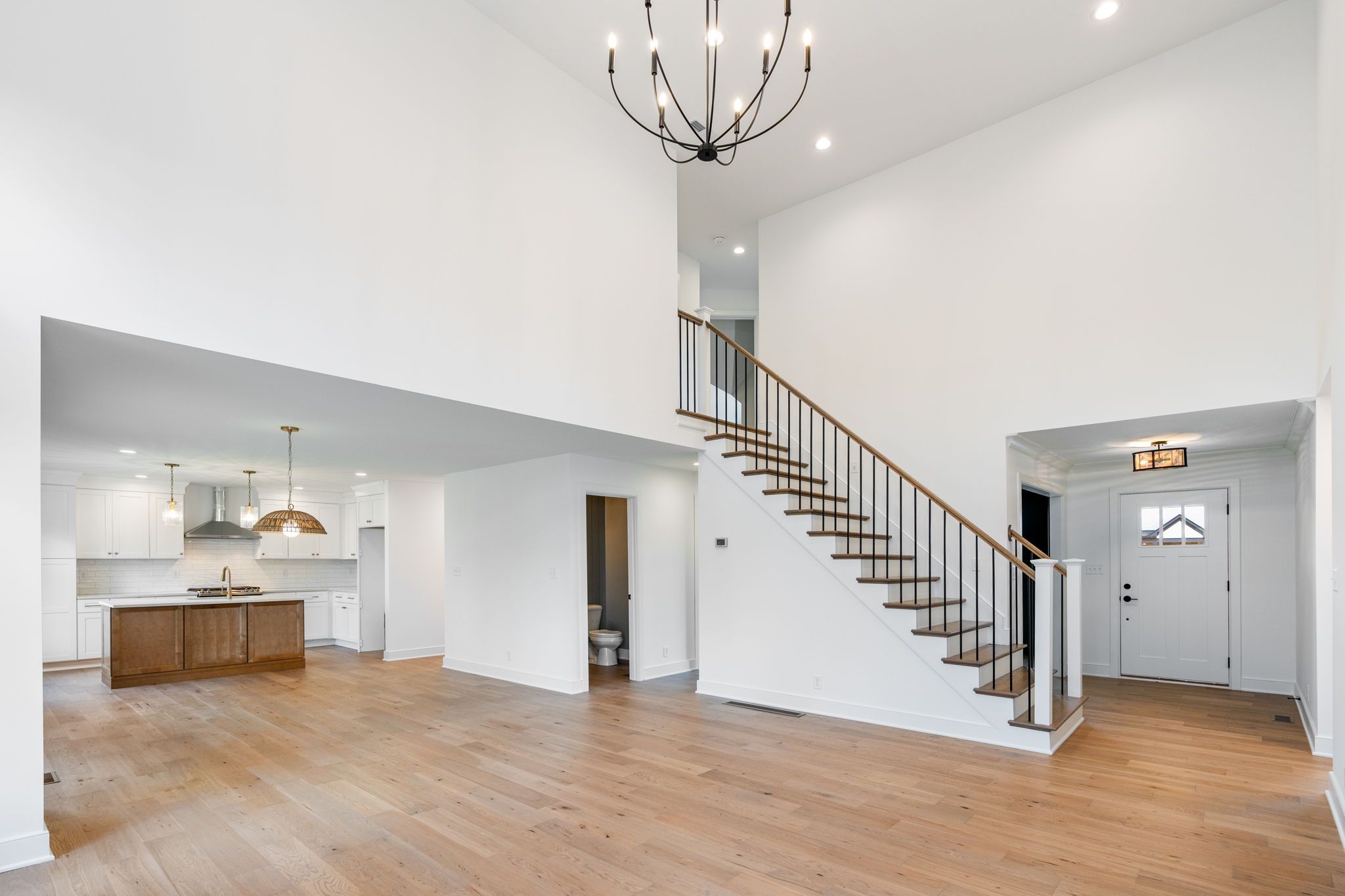 Two-story open living area with a staircase on the right, white walls, hardwood floors, and a black chandelier overhead.