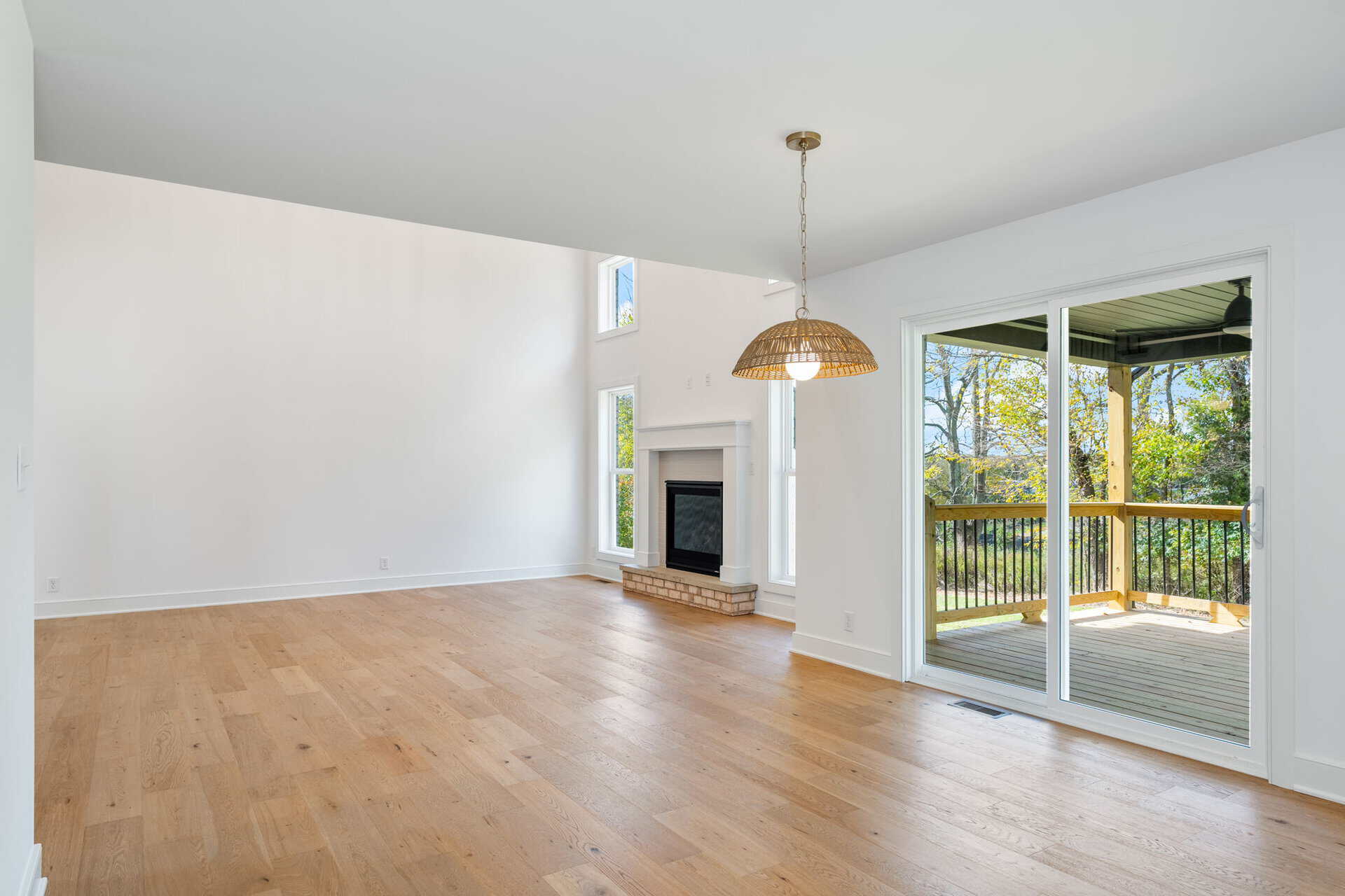 Bright living room with light wood flooring, brick fireplace, large windows, and a woven pendant light; sliding glass door to a wooden deck outside.