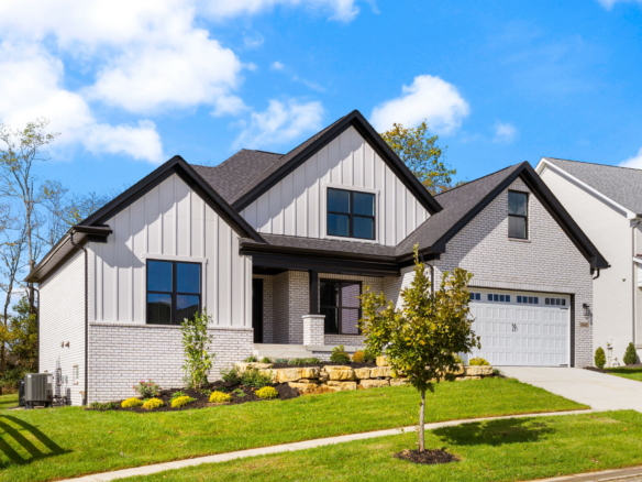 Two-story suburban house with brick and light siding, gabled roofs, and a manicured front yard under a blue sky.