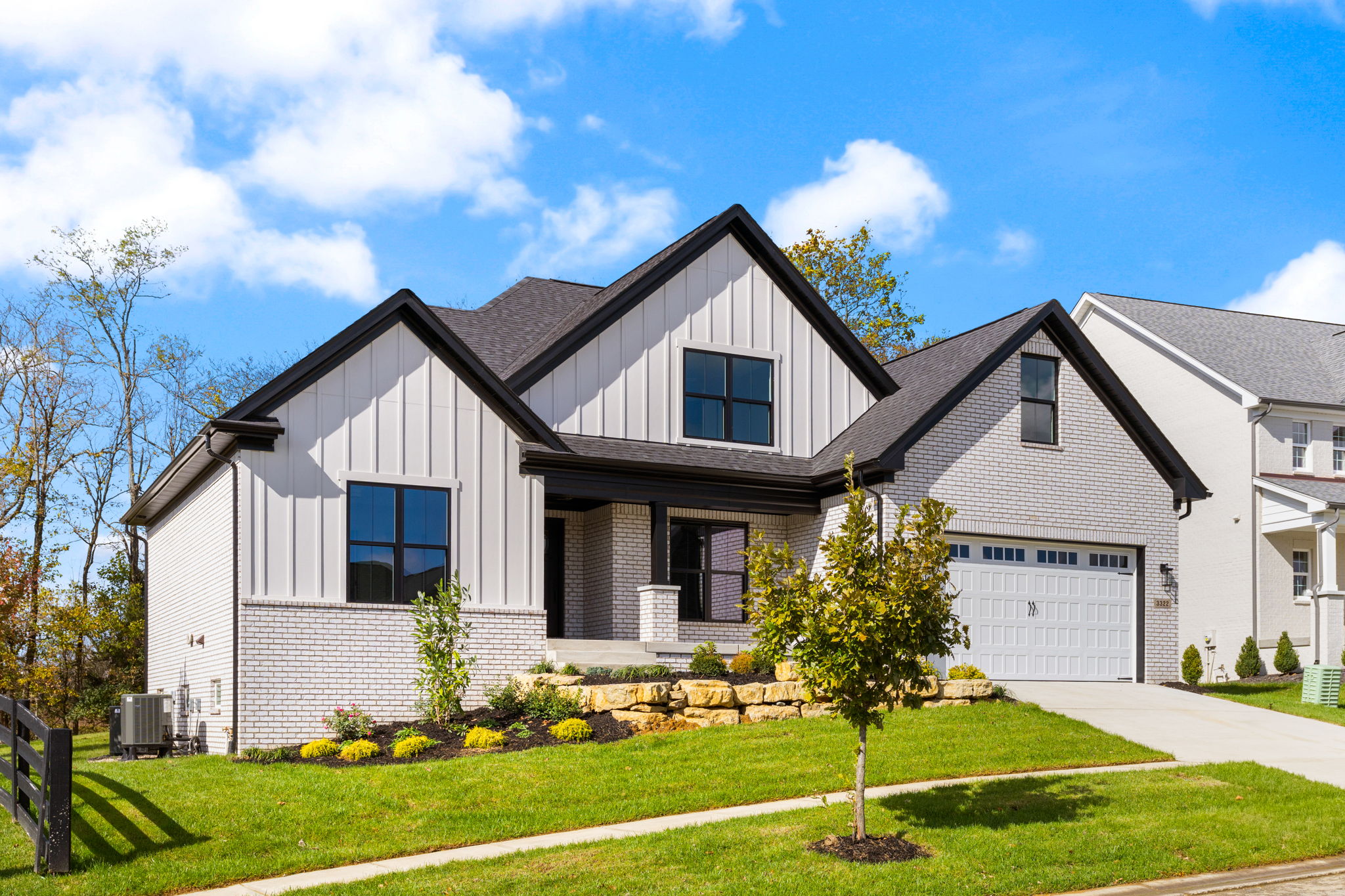 Two-story suburban house with brick and light siding, gabled roofs, and a manicured front yard under a blue sky.