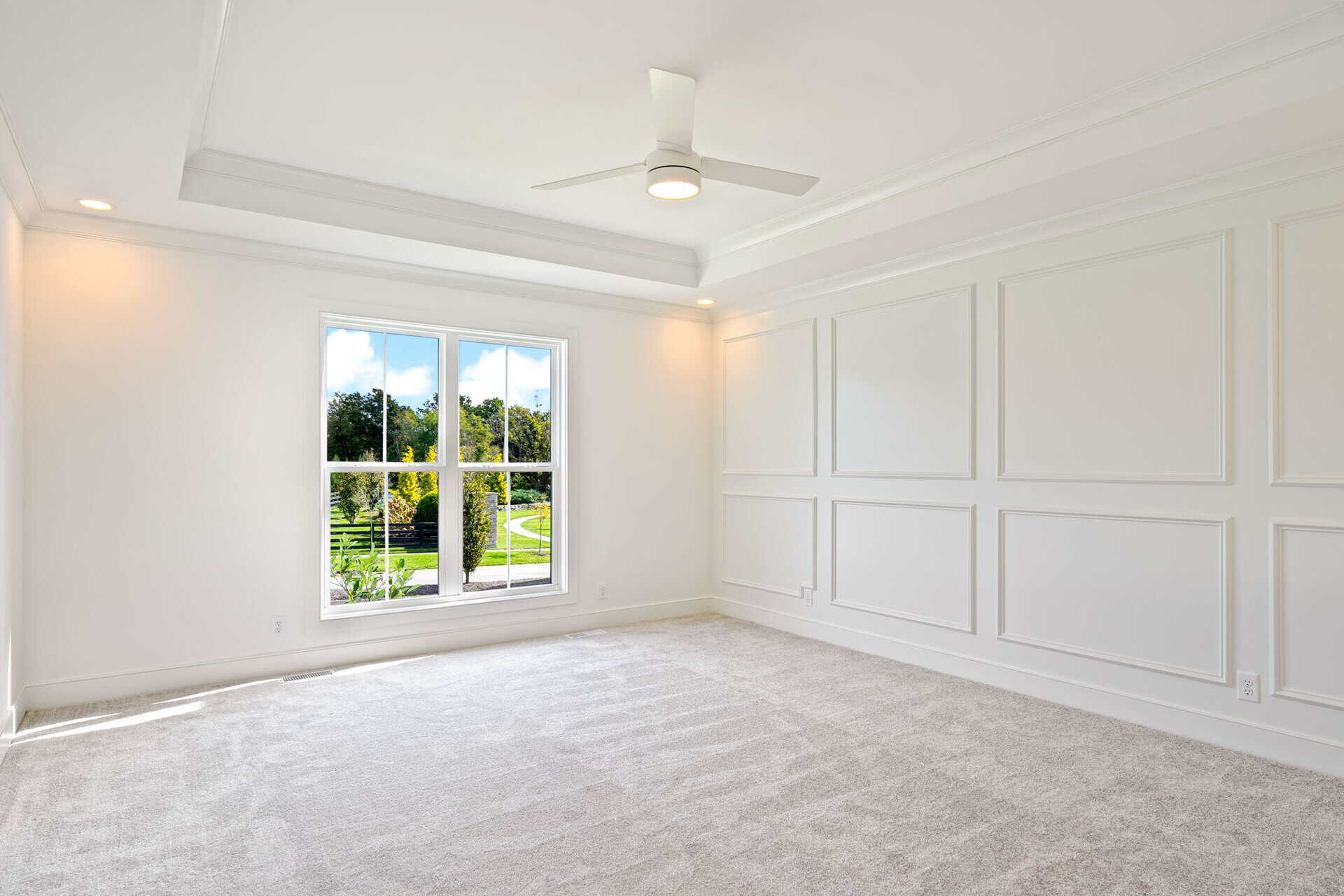 Bright empty room with white paneled walls, beige carpet, and a ceiling fan; a large window reveals a green yard outside.