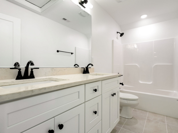 Bright bathroom with a double vanity, marble countertop, and black faucet fixtures reflected in a large mirror. A white tub/shower combo is visible in the background, plus a toilet nearby.