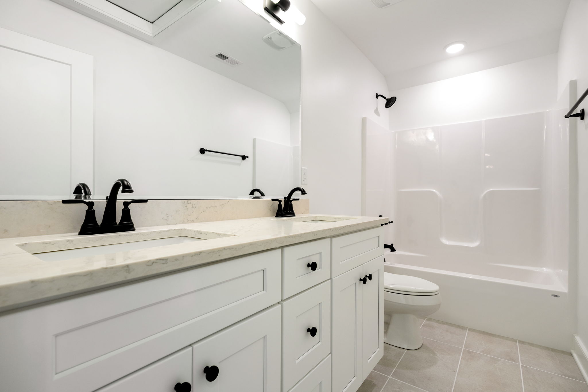 Bright bathroom with a double vanity, marble countertop, and black faucet fixtures reflected in a large mirror. A white tub/shower combo is visible in the background, plus a toilet nearby.