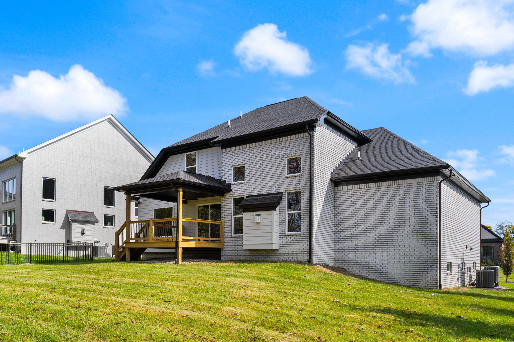 White brick suburban home with a wooden deck, large windows, and a green lawn under a bright blue sky with a few clouds.