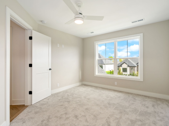 Empty bedroom with beige walls, white trim, and a large window revealing blue sky and suburban houses outside.
