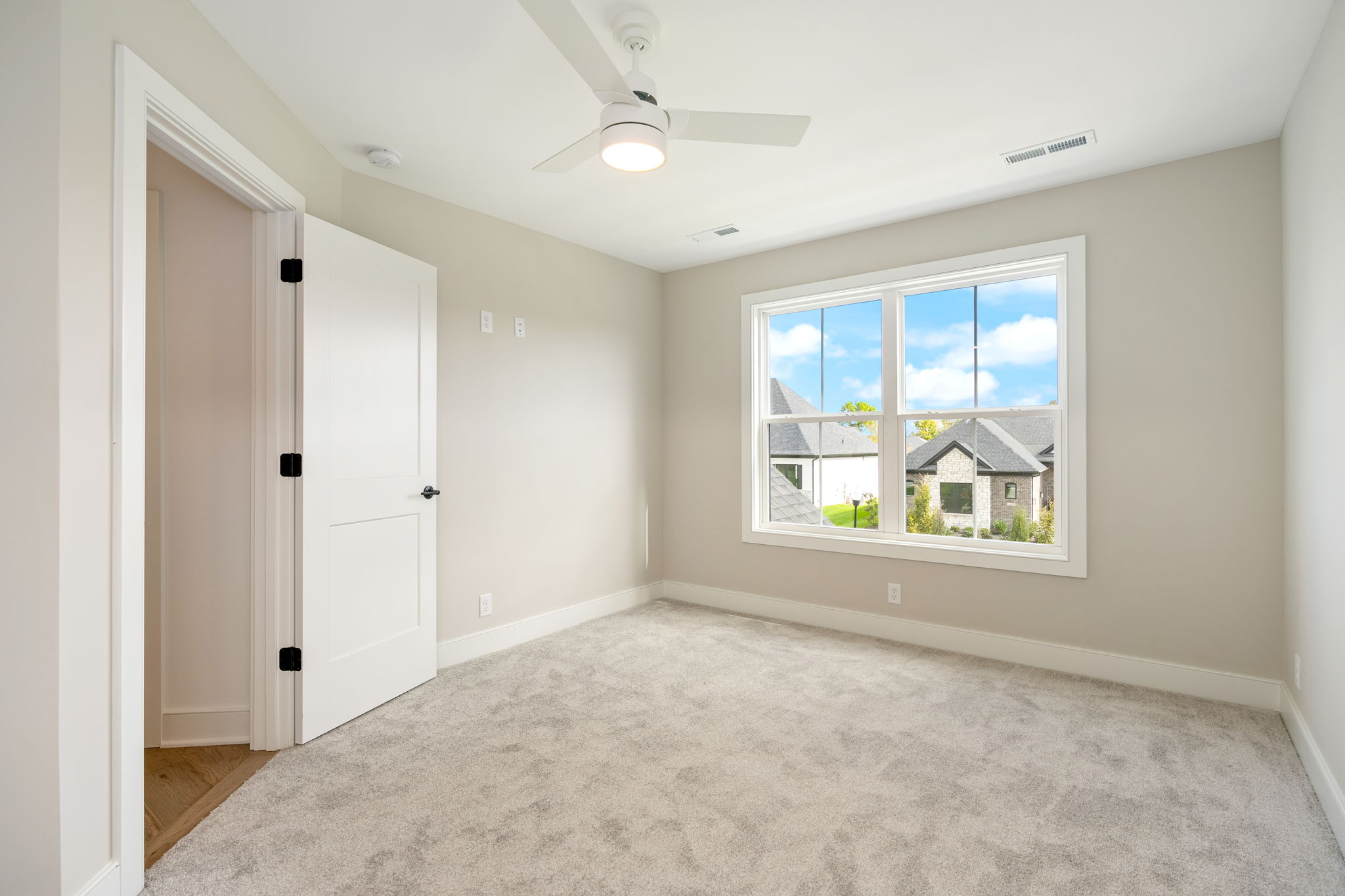 Empty bedroom with beige walls, white trim, and a large window revealing blue sky and suburban houses outside.