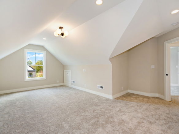 Sunlit empty attic room with beige walls, sloped ceiling, window overlooking a yard, and carpeted floor