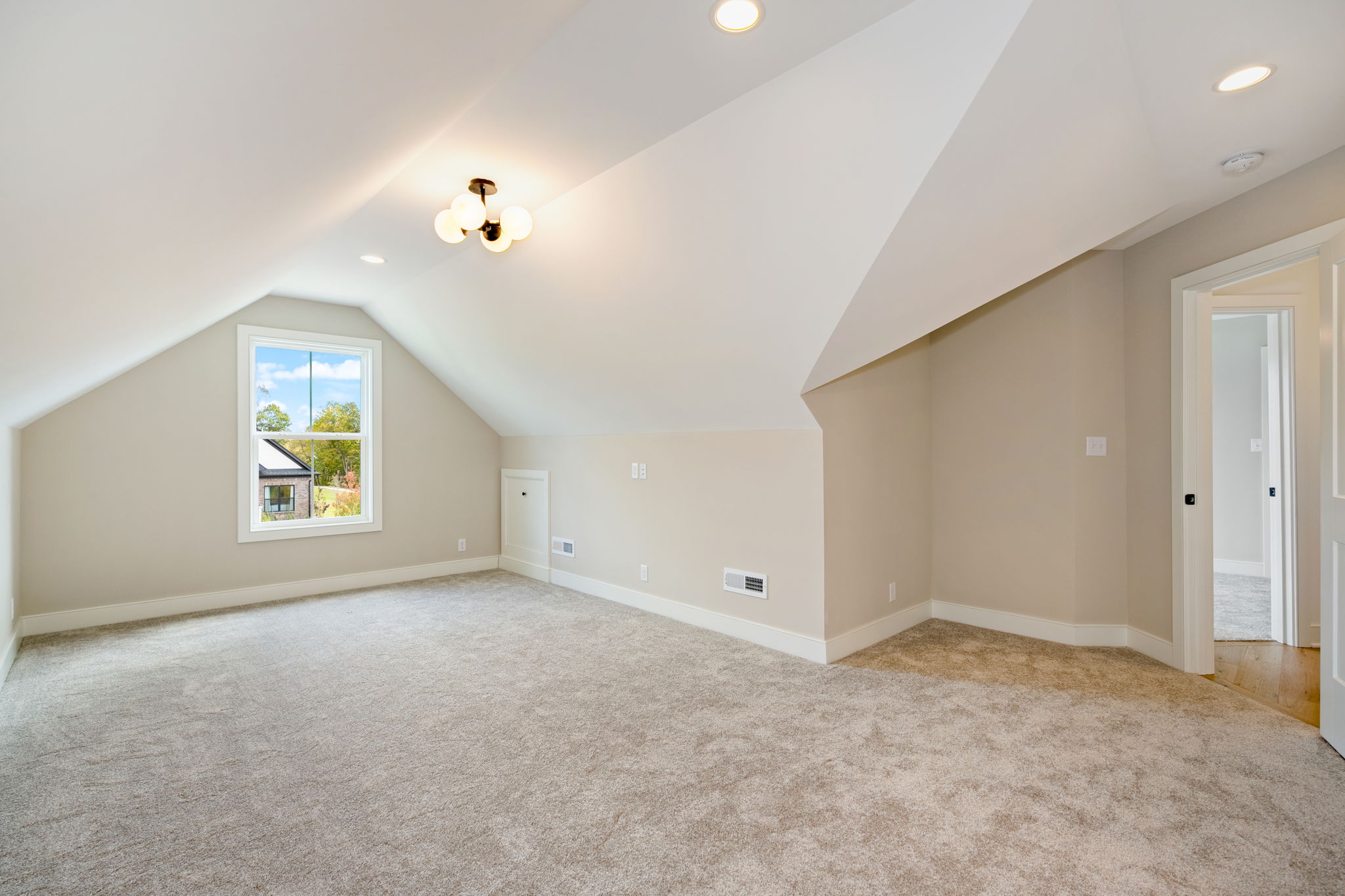 Sunlit empty attic room with beige walls, sloped ceiling, window overlooking a yard, and carpeted floor