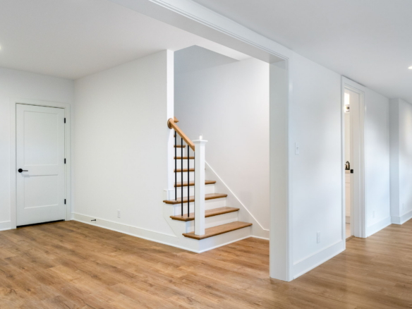 Bright hallway in a modern home with light wood floors, white walls, and a wooden staircase with black balusters at the center.