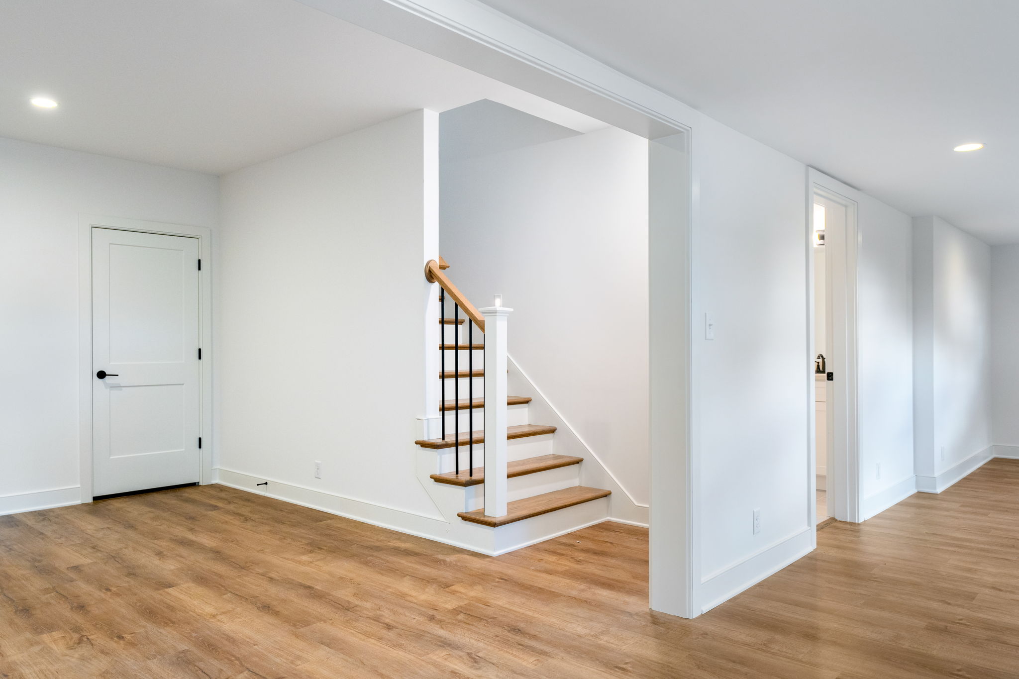 Bright hallway in a modern home with light wood floors, white walls, and a wooden staircase with black balusters at the center.