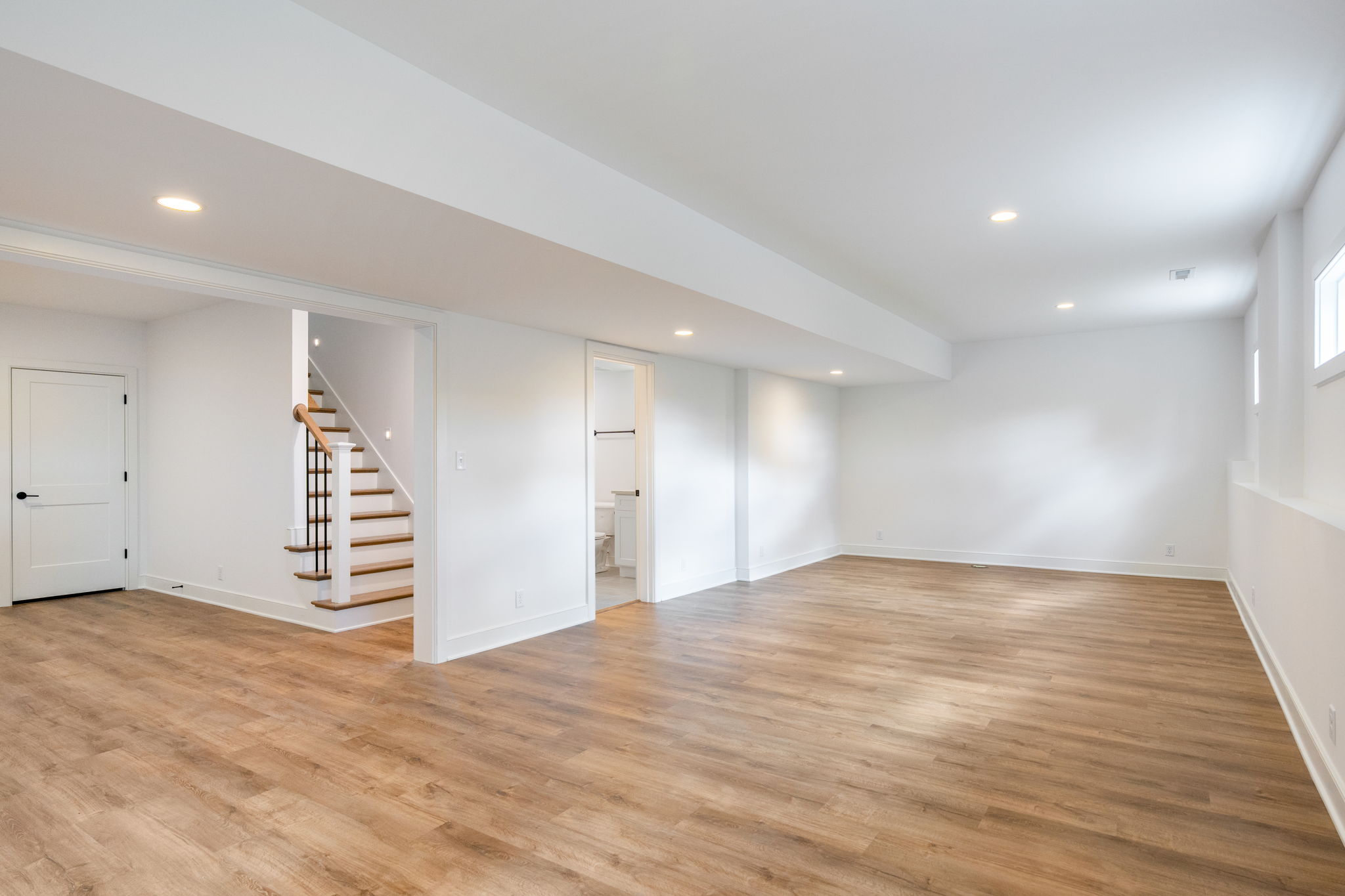 Empty finished basement with white walls, wood-look flooring, recessed lights, and a staircase on the left. Doors to rooms are visible along the back wall.