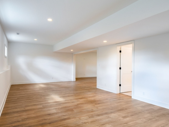 Empty finished basement with white walls and wood flooring, recessed ceiling lights, and an open doorway to another room.