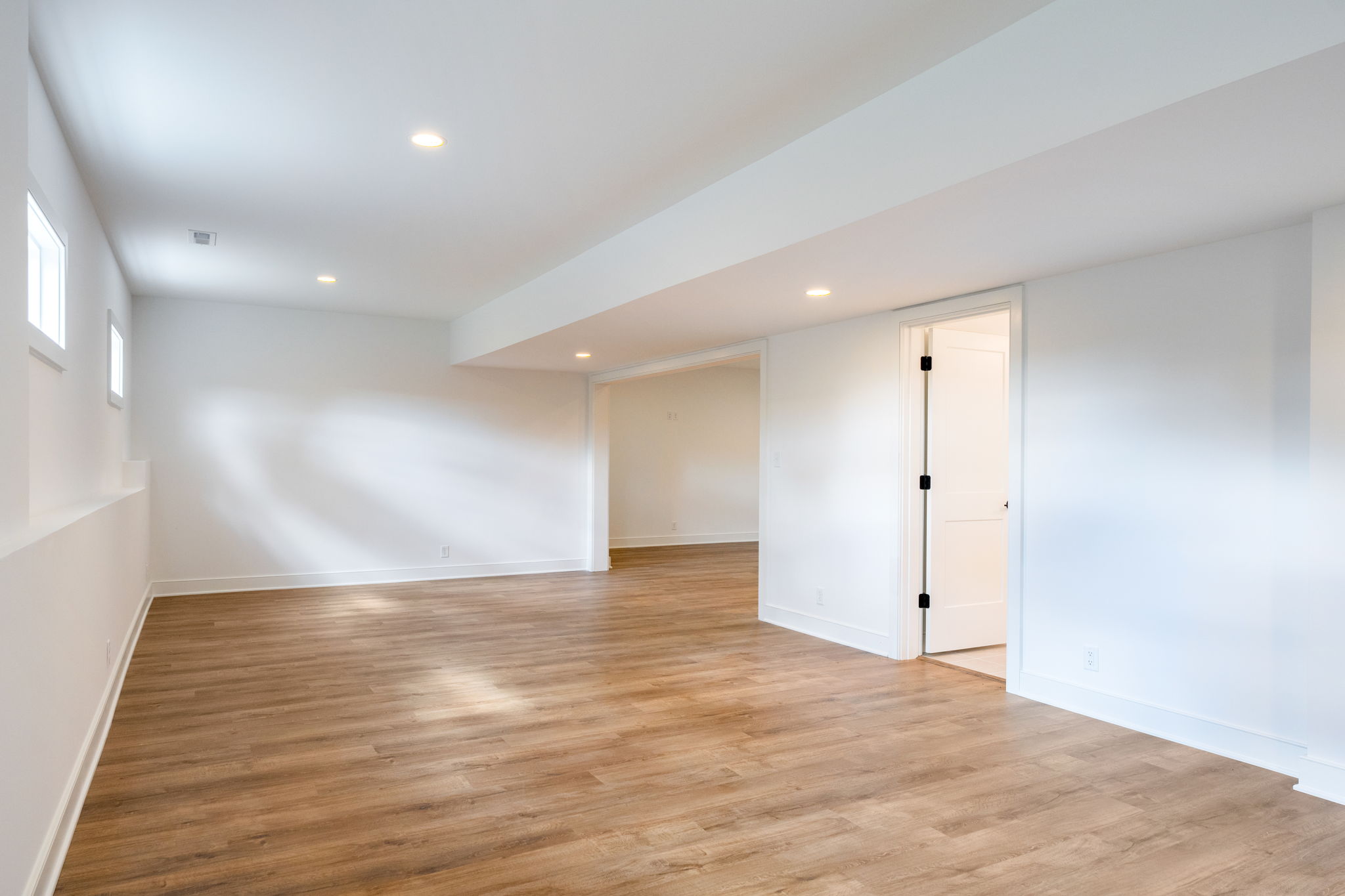 Empty finished basement with white walls and wood flooring, recessed ceiling lights, and an open doorway to another room.