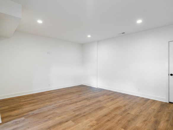 Empty white room with wood-tone flooring, recessed ceiling lights, and a white door on the right side.