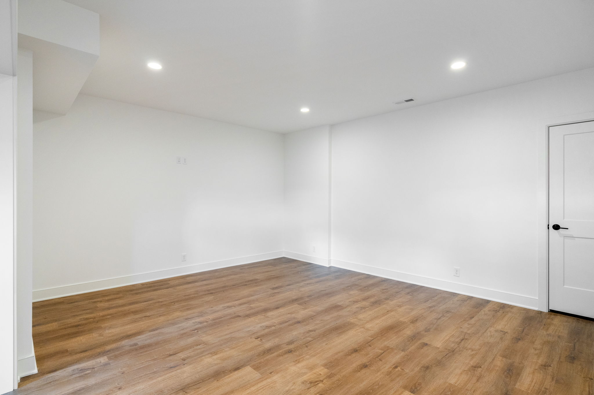 Empty white room with wood-tone flooring, recessed ceiling lights, and a white door on the right side.