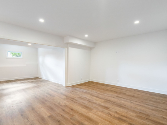 Bright empty room with white walls, wood-look flooring, recessed ceiling lights, and a small horizontal window.