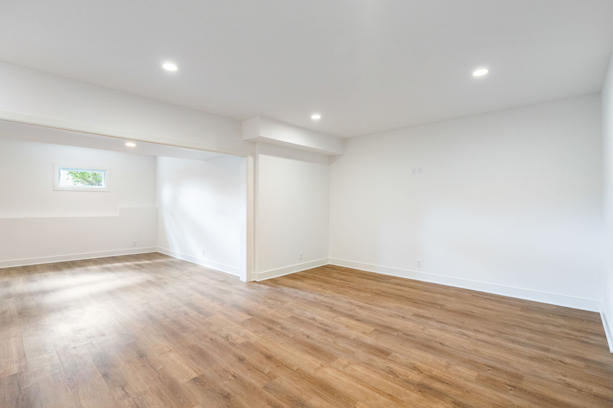 Bright empty room with white walls, wood-look flooring, recessed ceiling lights, and a small horizontal window.