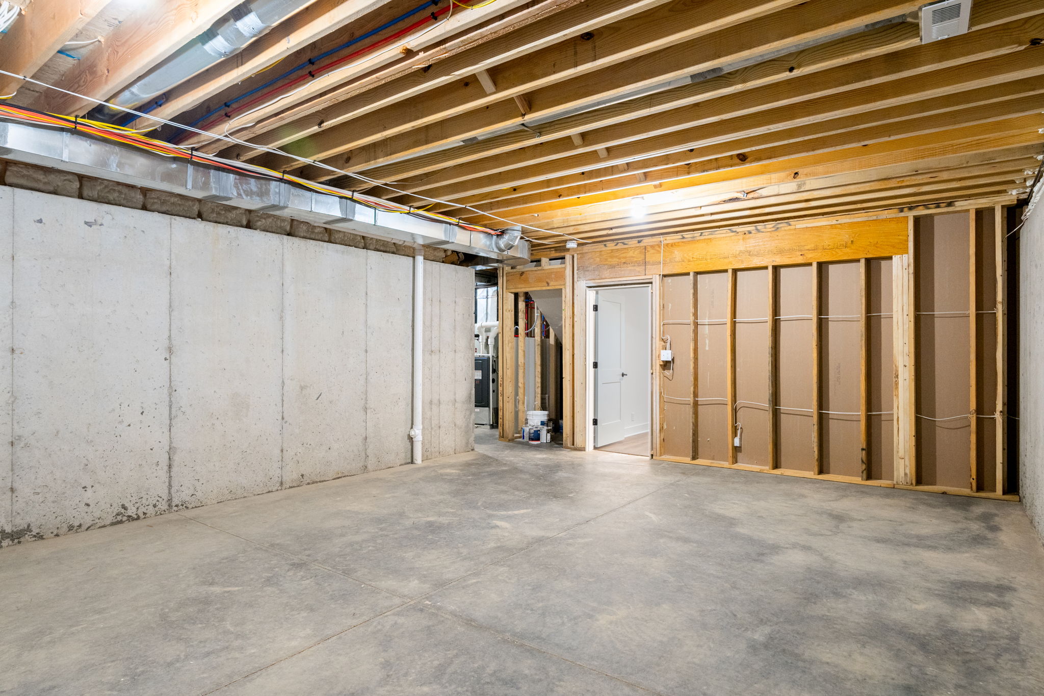 Unfinished basement with exposed joists, concrete walls, and visible wiring; doorway to a back room in renovation area.