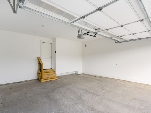 Empty residential garage with a white door at back and a ceiling-mounted metal track garage door system; small wooden stairs on left.