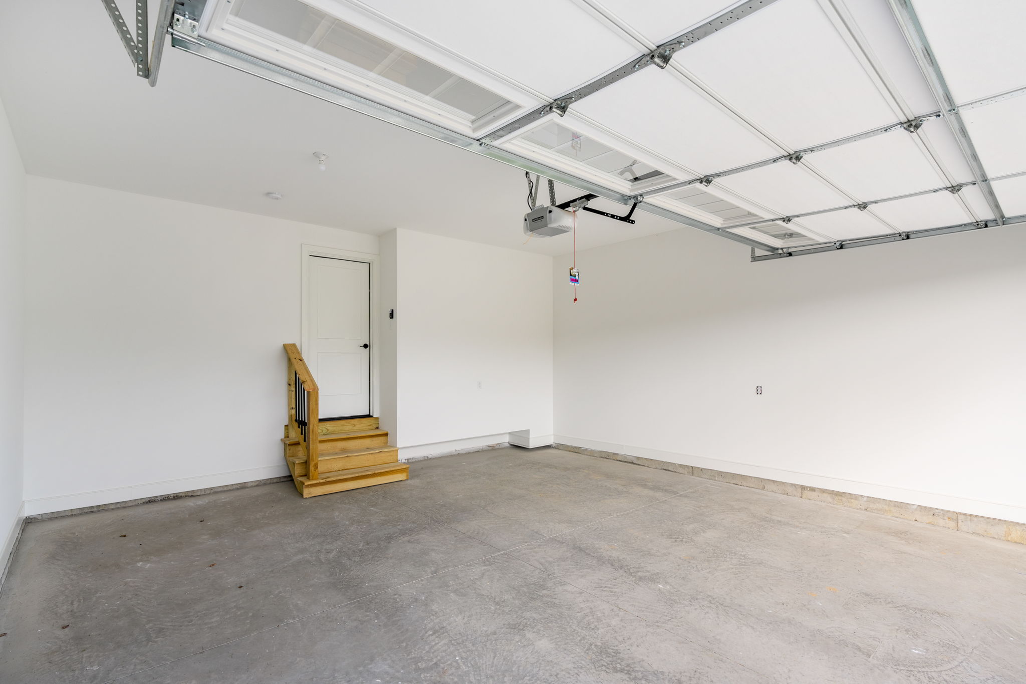 Empty residential garage with a white door at back and a ceiling-mounted metal track garage door system; small wooden stairs on left.
