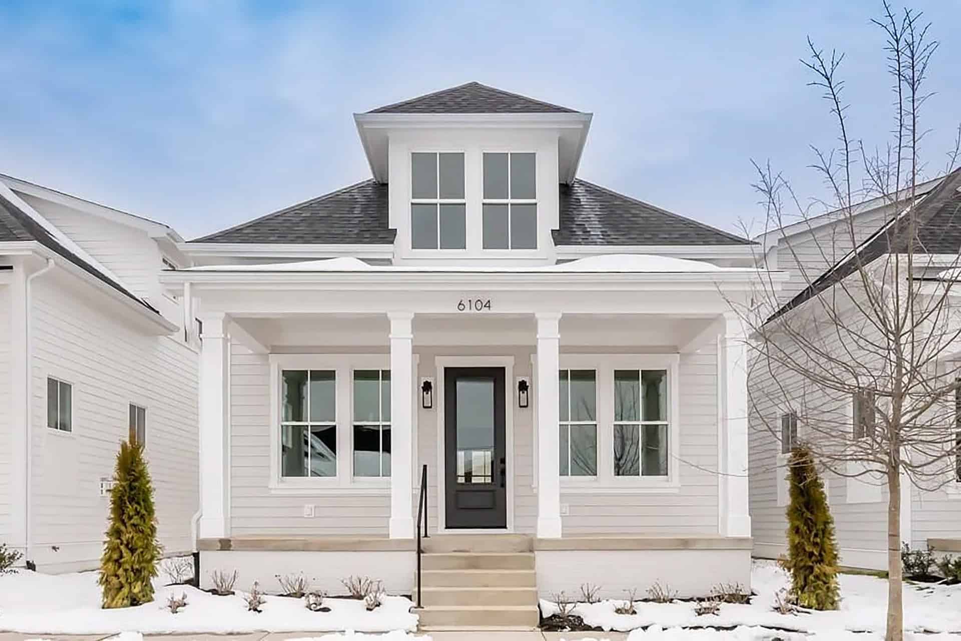 6104 Passionflower Drive Modern white house with black door and dormer window, snow on ground.