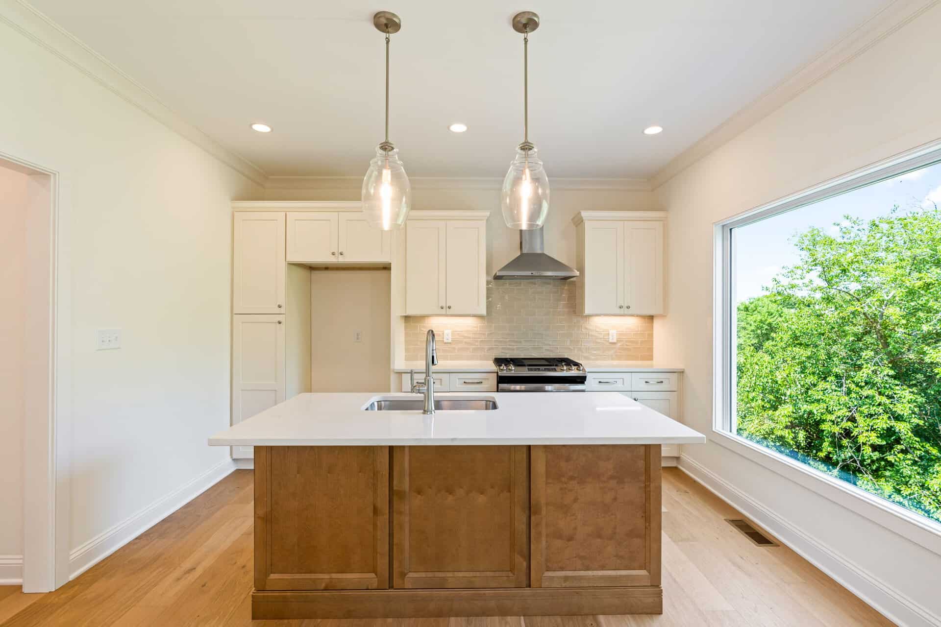 Bright kitchen with white cabinets, stainless steel range, and a wooden island; two glass pendant lights and a large window overlooking trees.