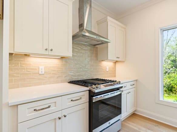 Modern kitchen with white cabinets, stainless steel stove, and large window overlooking greenery.