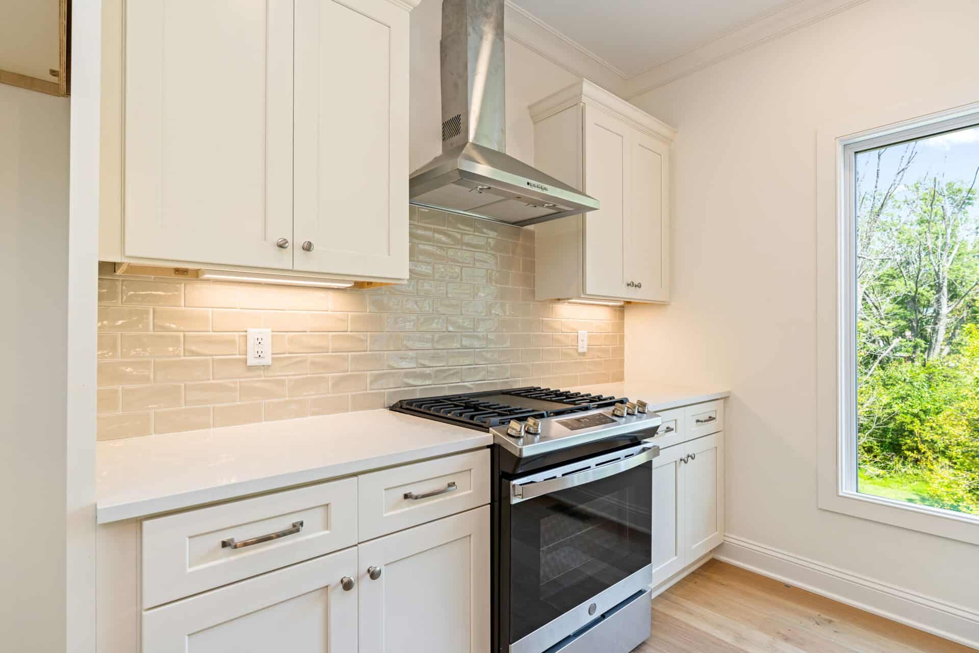 Modern kitchen with white cabinets, stainless steel stove, and large window overlooking greenery.