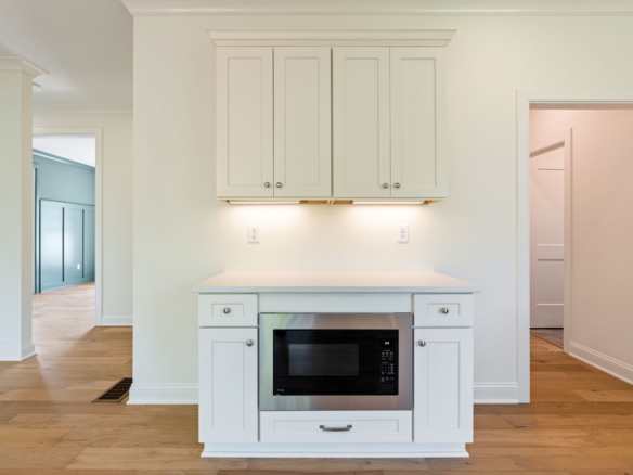 White kitchen island with built-in microwave, oven, and upper cabinets against a light wall; warm hardwood floors illuminate the space.