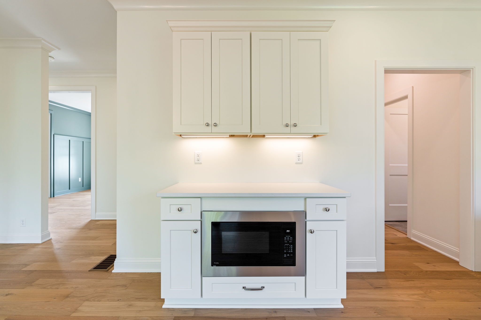 White kitchen island with built-in microwave, oven, and upper cabinets against a light wall; warm hardwood floors illuminate the space.