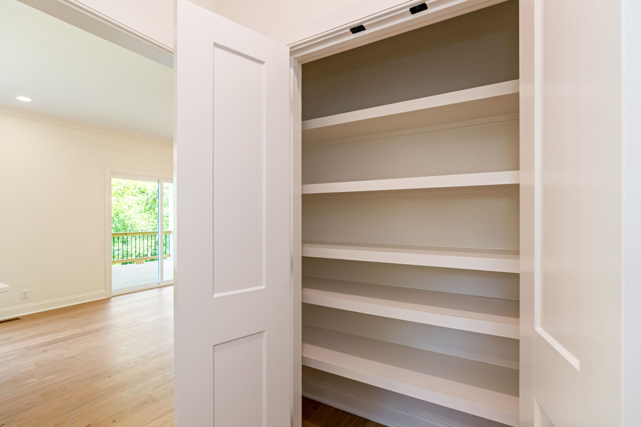 Open white built-in closet with multiple empty shelves, doors swung to the side in a bright room with wood floor