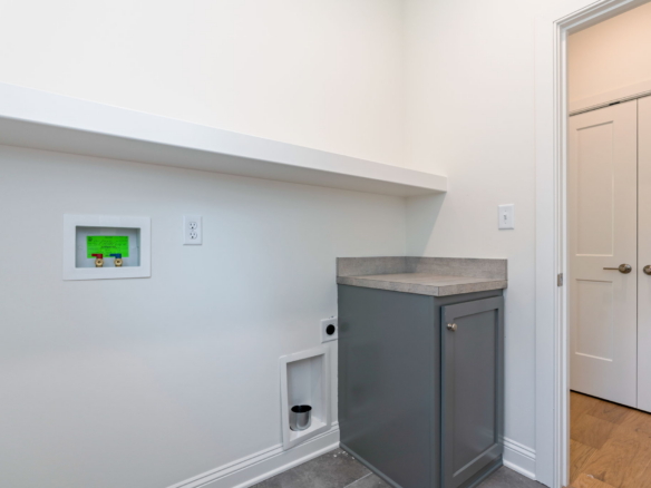 White-walled utility nook with a gray base cabinet and light gray countertop, small recessed utility box with green label, and a doorway with double closet doors on the right.