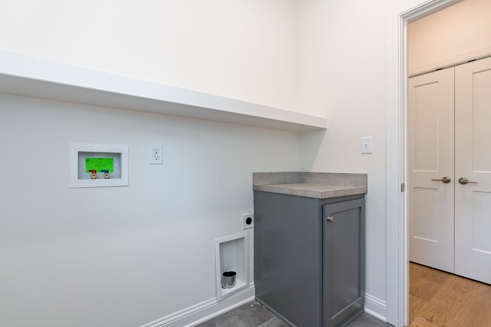 White-walled utility nook with a gray base cabinet and light gray countertop, small recessed utility box with green label, and a doorway with double closet doors on the right.