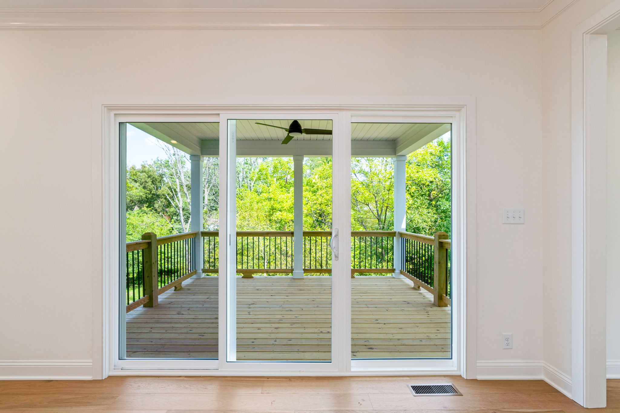Sliding glass door opening to a wooden deck with railing; ceiling fan overhead and green trees visible outside.