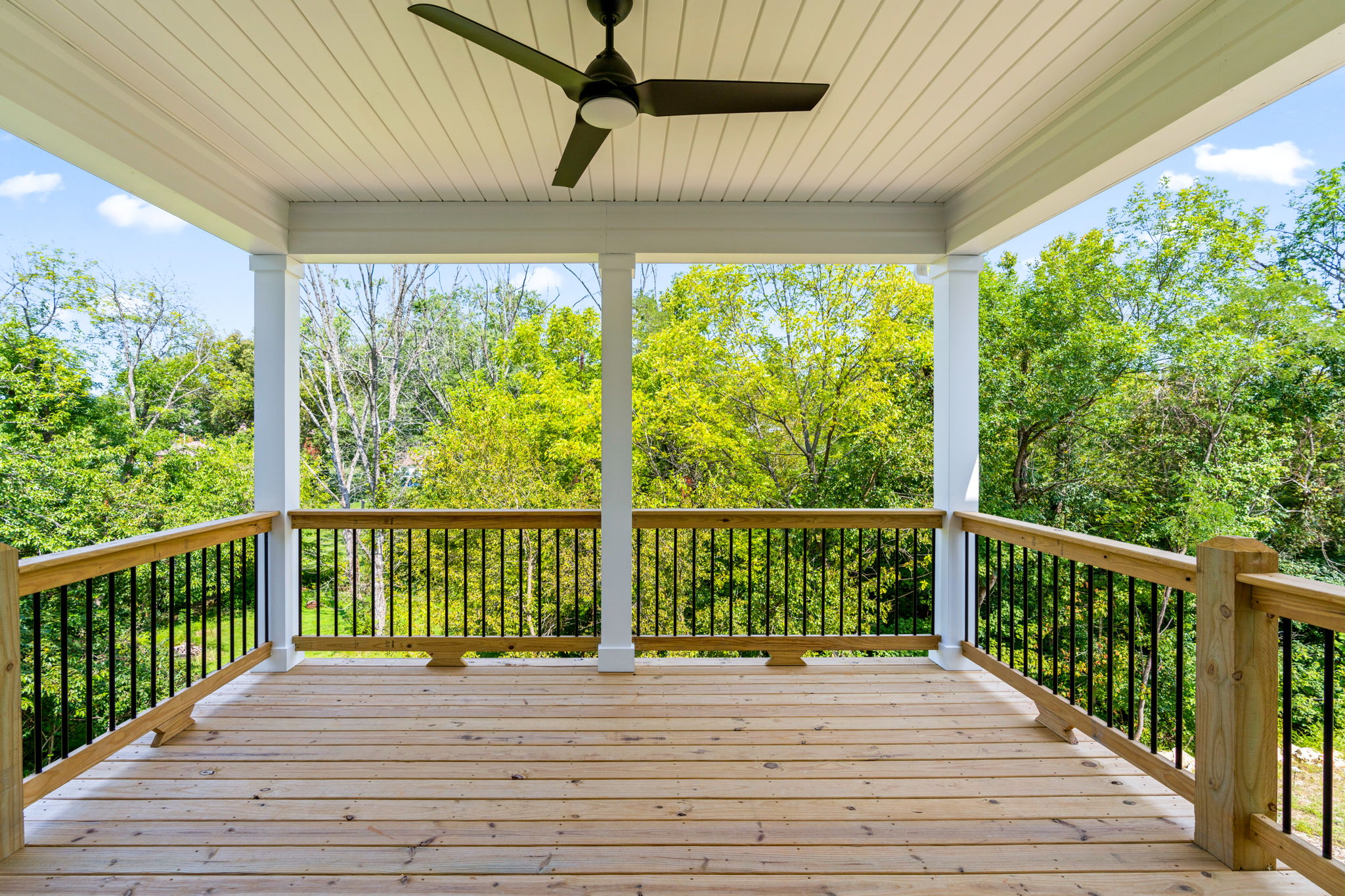 Open-air wooden deck with a ceiling fan overhead, white posts, and black railing, overlooking a lush green treeline.