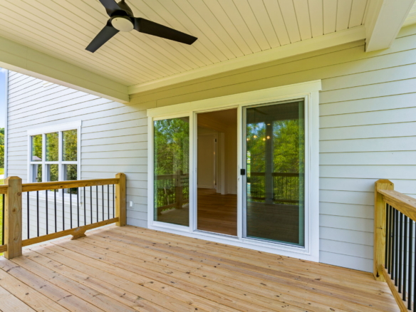 Wooden deck attached to a house with sliding glass doors, beige siding, and a ceiling fan overhead, overlooking a green yard.