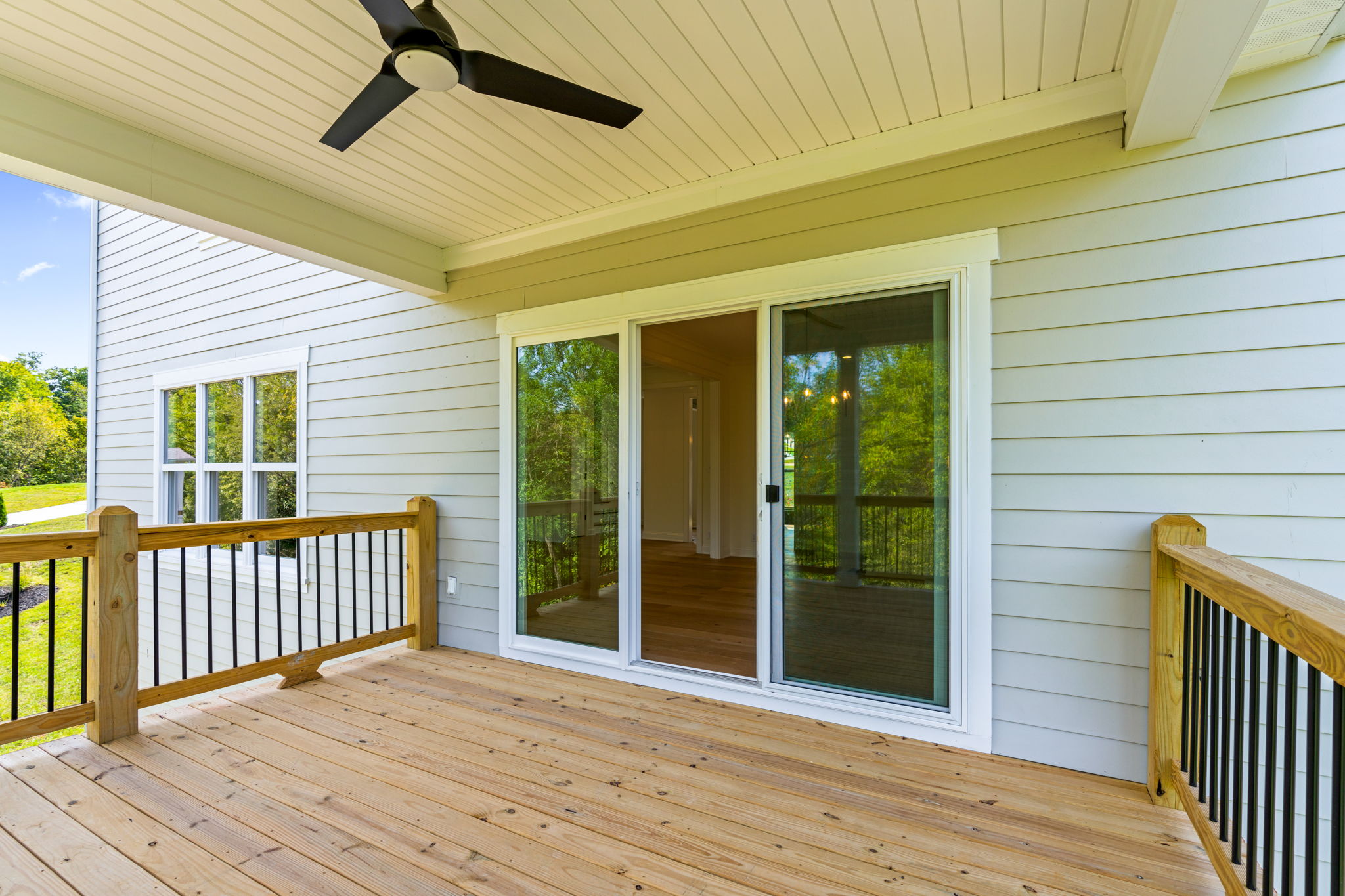 Wooden deck attached to a house with sliding glass doors, beige siding, and a ceiling fan overhead, overlooking a green yard.