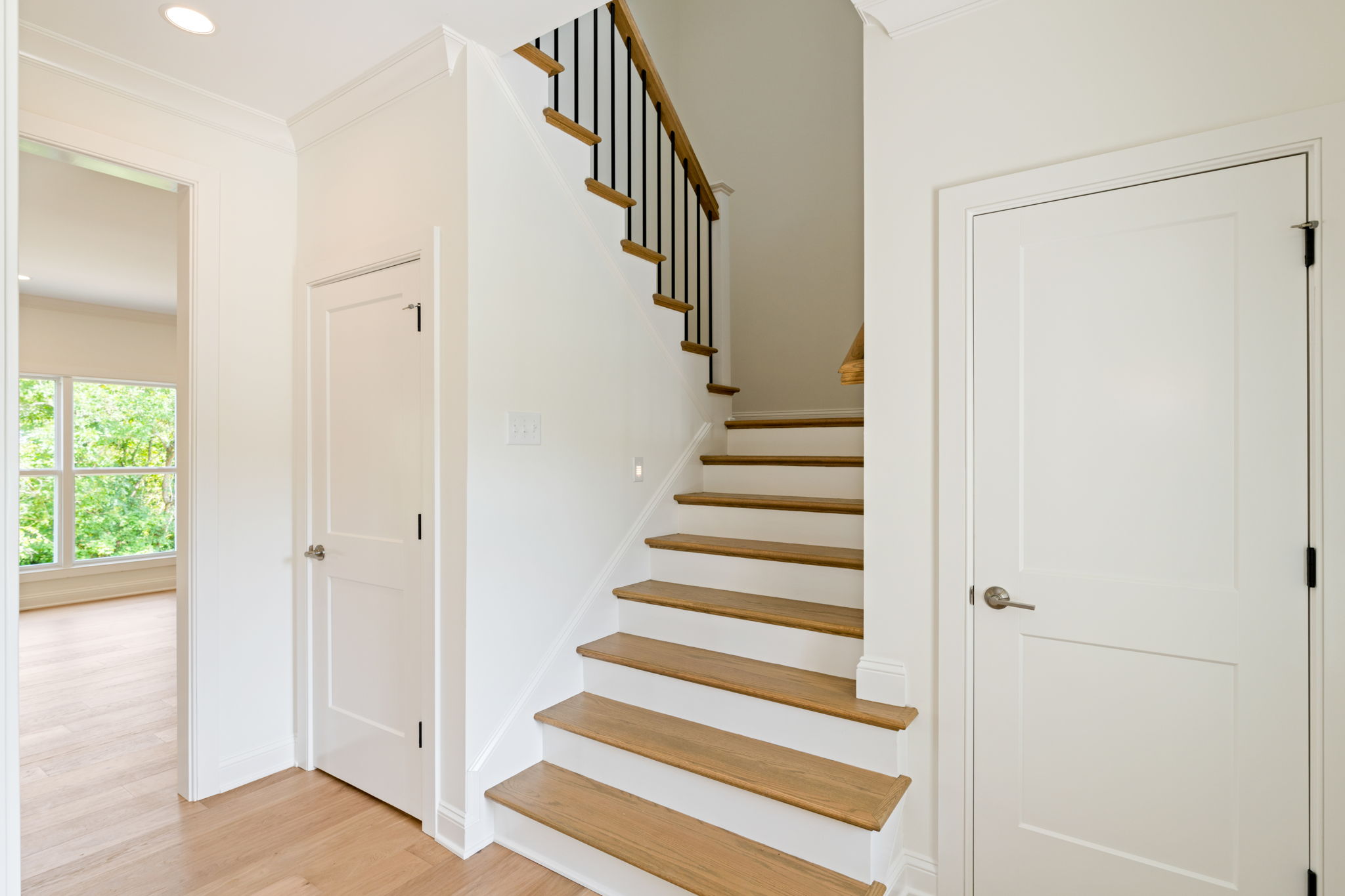 Interior staircase with wooden treads and black metal balusters along white walls, in a bright hallway with doors nearby
