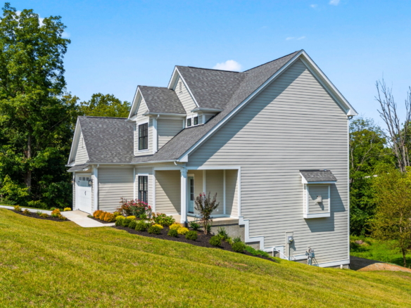 Front view of a two-story beige house with multiple gabled roofs and white trim on a sunny day, with green lawn and trees nearby.